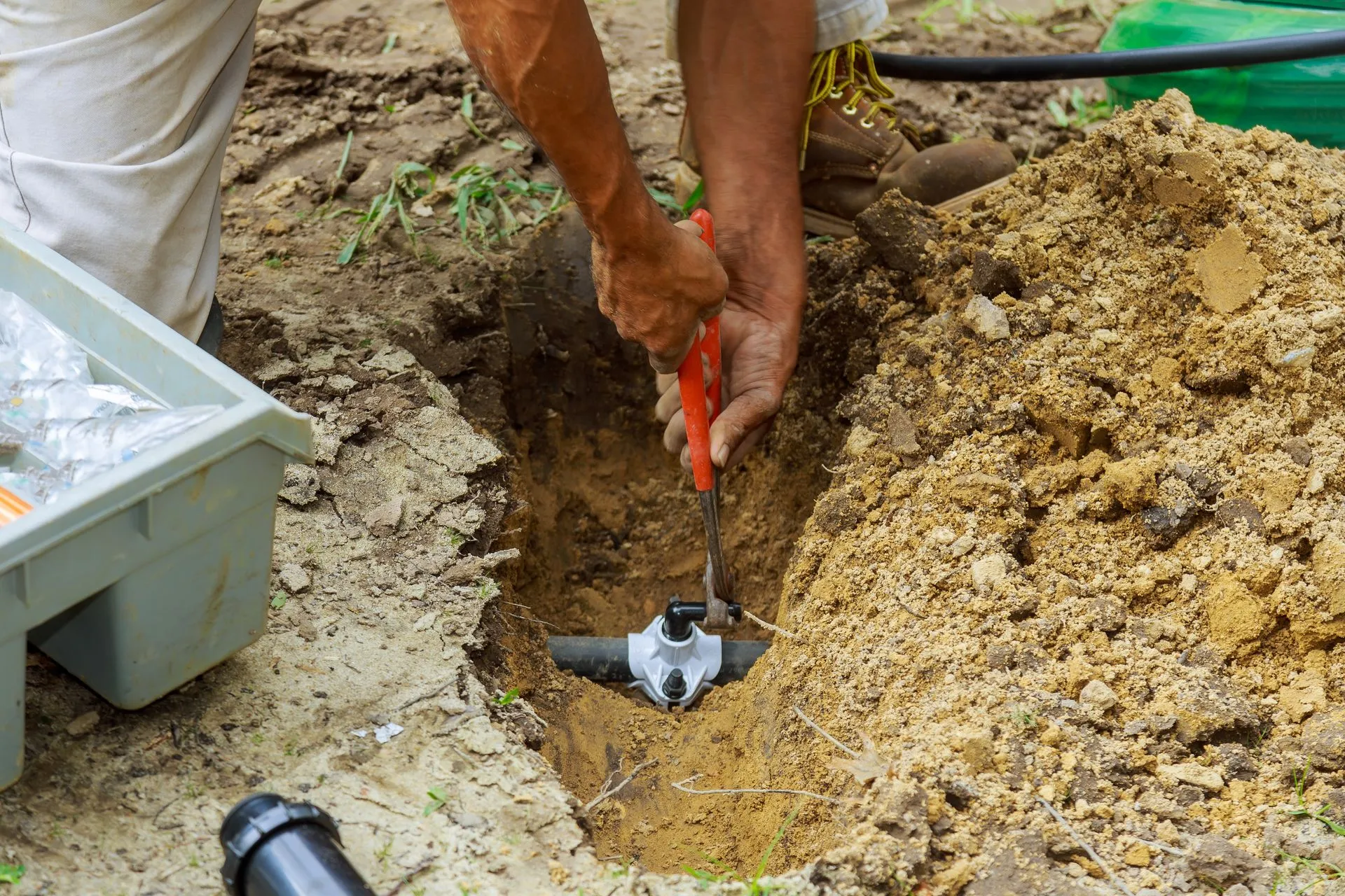 Person using pliers on a pipe fitting in a hole in the ground. A toolbox sits nearby.
