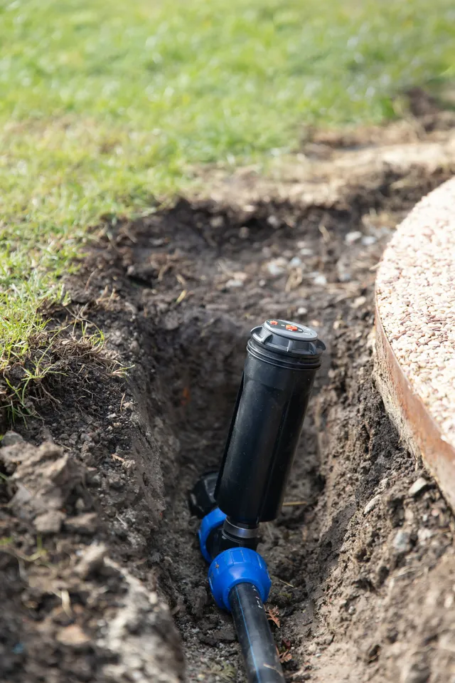 Sprinkler head in a trench, connected to a pipe, near a lawn and walkway.