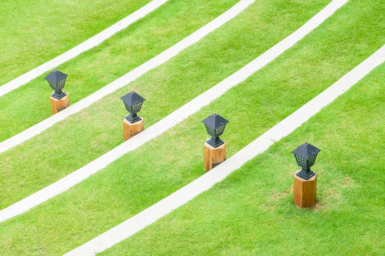 Four black outdoor lamps on wooden posts stand spaced out in a row on a lawn with curved white paved pathways.