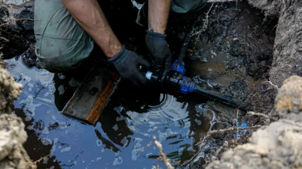 A worker in protective gloves repairing a black plastic water pipe in a muddy, water-filled excavation trench.